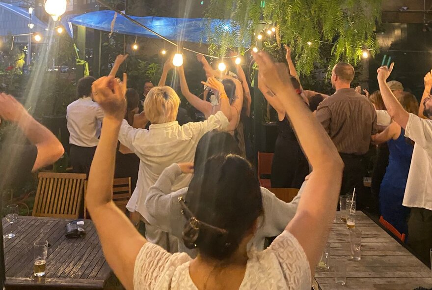 People in an outdoor setting with fairy lights raising their hands in the air.