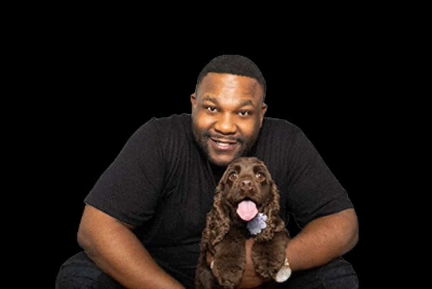 Comedian Nate Jackson smiling and wearing a black t-shirt, a brown dog sits in front of him.