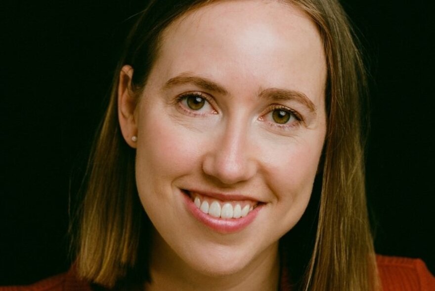 Young woman with a open mouthed smile showing her teeth, and straight brown hair, against a dark background.