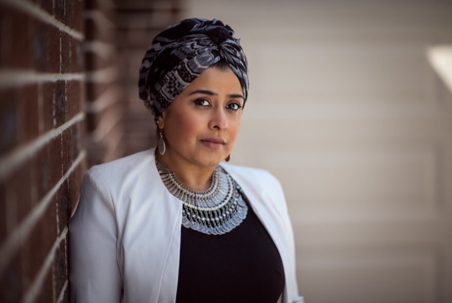 Woman wearing a turban and heavy silver jewellery, standing next to a brick wall.