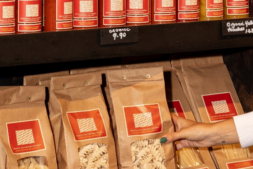 Rows of authentic brown paper pasta packets on a shelf; a woman's hand picking one up.