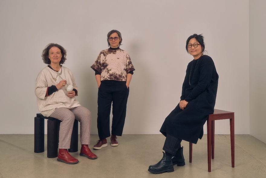 Three people in a gallery space, two seated on seats, one standing with hands in pockets next to a wall.