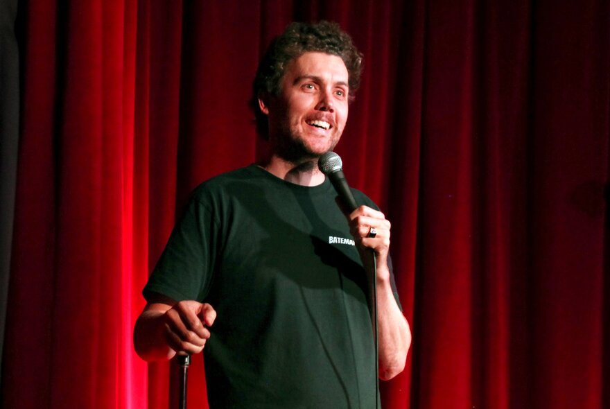 A comedian talking into a microphone they hold in their hand, while standing on a stage in front of a red curtain.