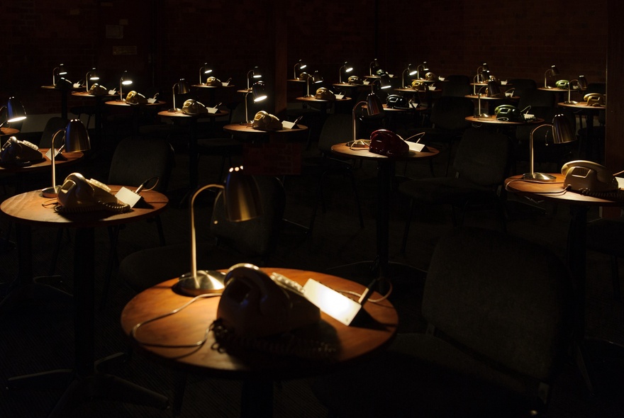 Multiple small tables of vintage phones illuminated by desk lamps, part of an interactive theatre installation at the Malthouse Theatre.