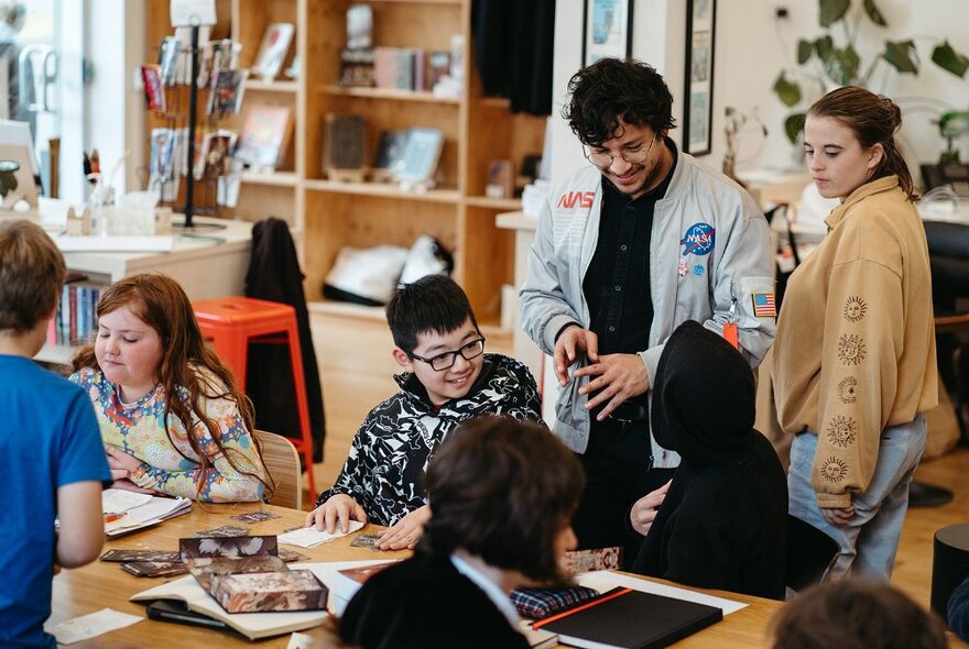 Several teenagers sitting and working around a large table in a room, with their adult teacher interacting with them, bookshelves in the background.