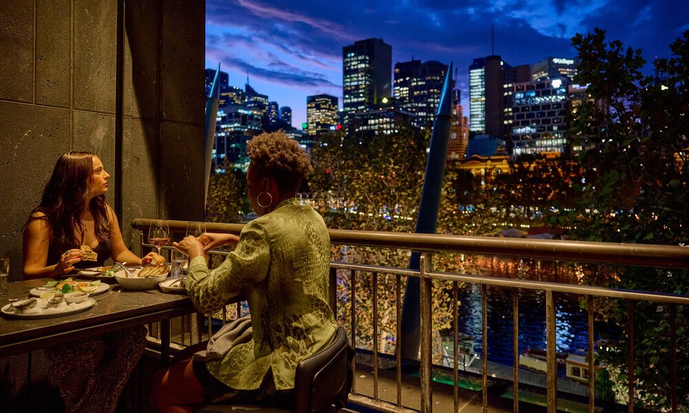 Two friends are eating dinner on a balcony at a restaurant at dusk. There is a view of the Yarra river and the city skyline.