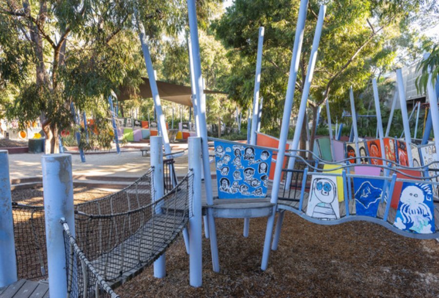 Pale blue playground with art murals, a bridge and trees.