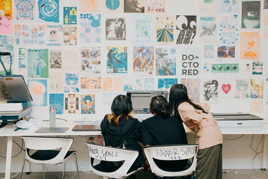 Three students seated around a computer monitor at a workbench, the studio wall in front of them filled with posters and designs.