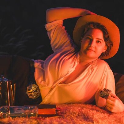 Comedian Laura Davis wearing a cowboy outfit while reclining next to a swag.