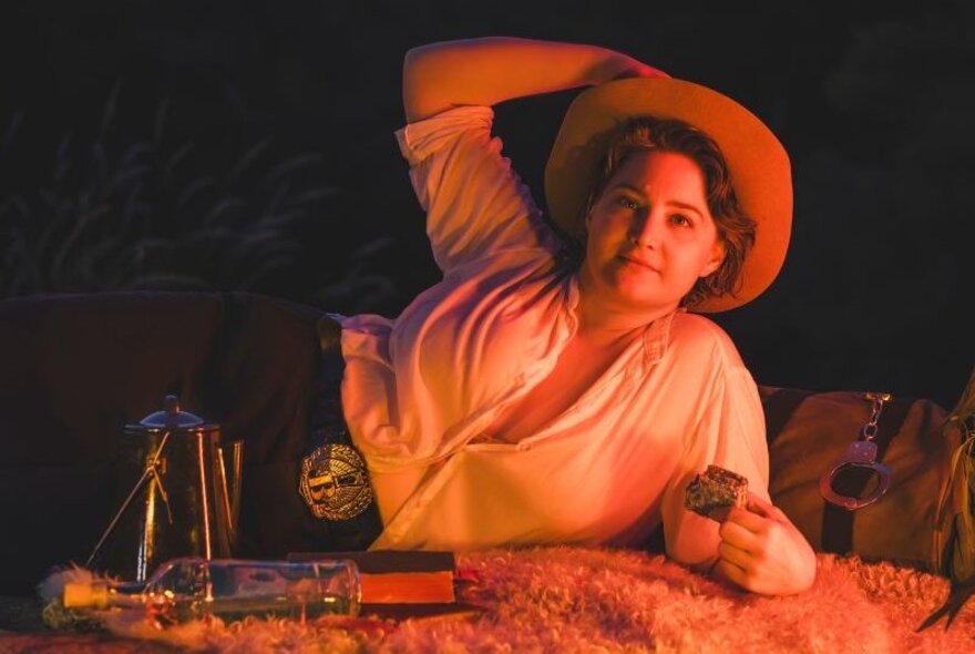 Comedian Laura Davis wearing a cowboy outfit while reclining next to a swag.