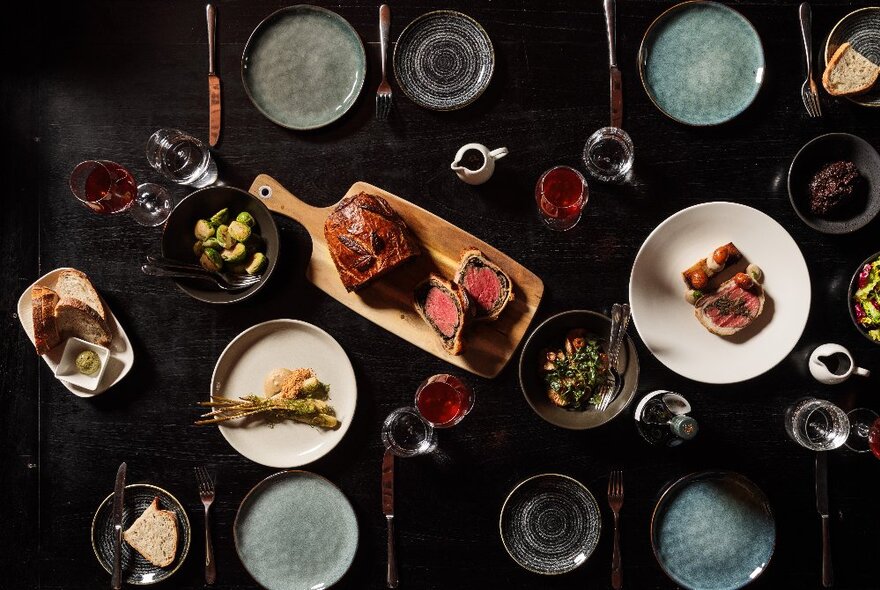 Christmas fare arranged on plates and displayed on a black surface with wine glasses, crockery and cutlery, seen from above. 