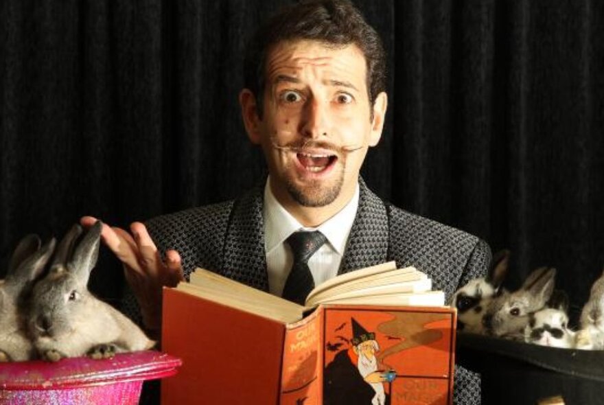 A magician looking slightly terrified while reading from an orange spell book with a rabbit to his right.