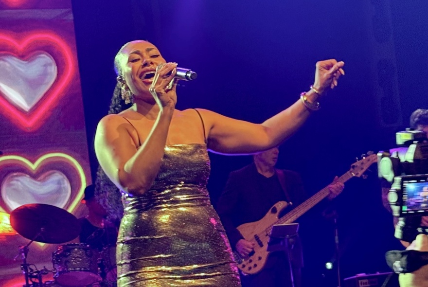 Singer Paulini on stage in a gold sequined dress, singing into a microphone.