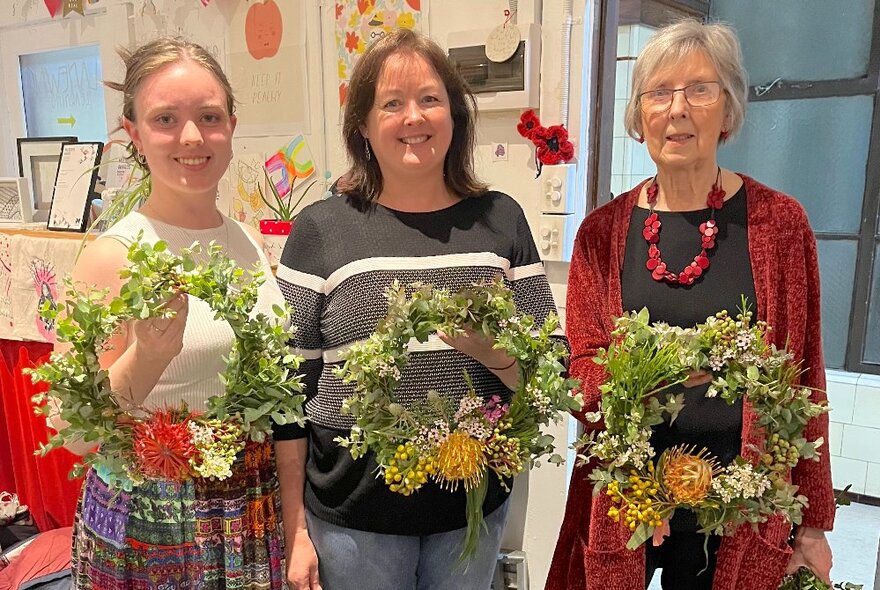 Three people in a studio holding up circular wreaths they've made using native flowers and greenery.