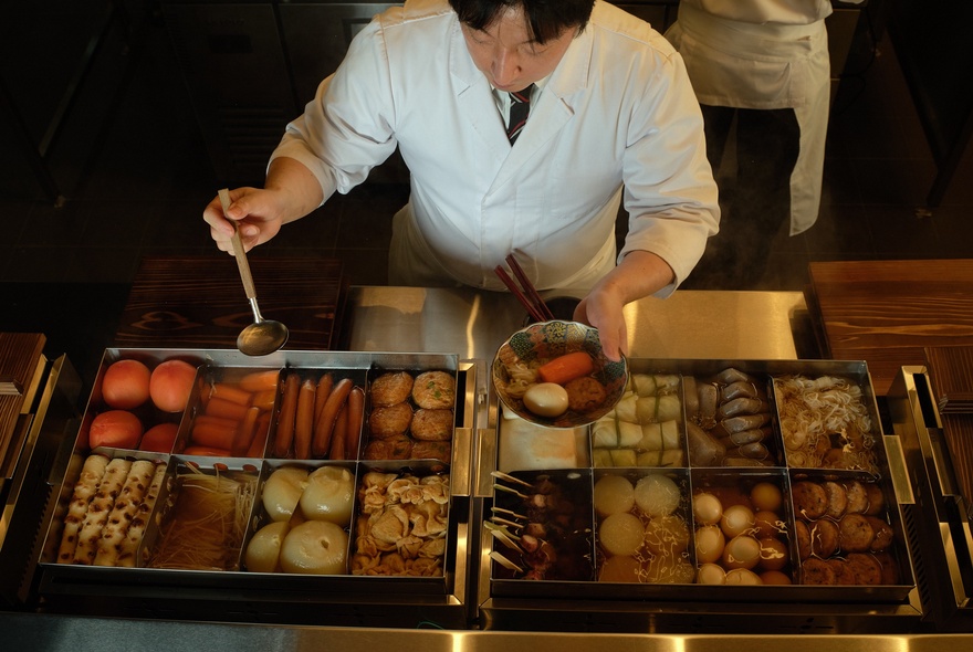 Japanese chef holding a bowl of food and spooning on ingredients from a large selection of dishes.