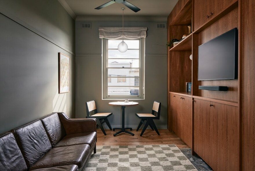 The sitting room with leather couch facing a TV unit in a suite at The Courtroom Hotel, a small table with two chairs near the window on the rear wall.