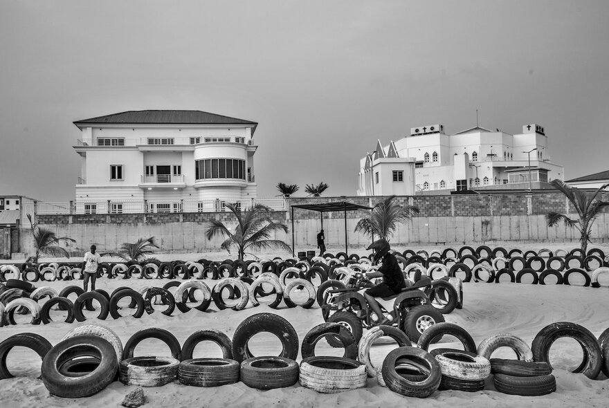 A black and white photo of a sandy beach area with numerous used car tyres arranged in rows, a person on a motorbike riding on the track, in the background are two large, modern white houses.