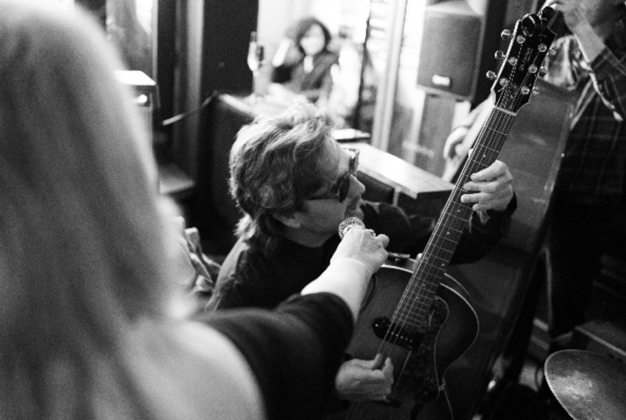 Black and white image of person playing an acoustic guitar while another individual holds a microphone for them.