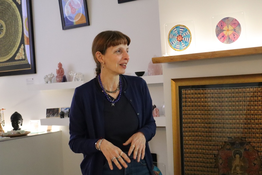 Woman in black, seated with hands on her knees, in a studio with ceramic artworks on the wall.