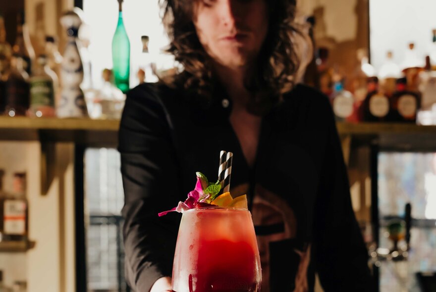 A bartender holding out a dark red cocktail with a pink garnish and two striped straws.