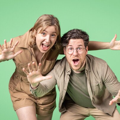 A man and a woman lean forward with wide, surprised facial expressions and open mouths, both extend their hands toward the camera with fingers spread wide in a playful, startled gesture. 