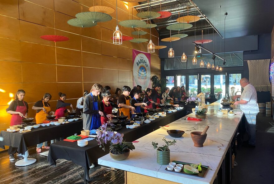 People working at long tables set up with cooking stations and ingredients and cooking utensils during a Thai cooking workshop.