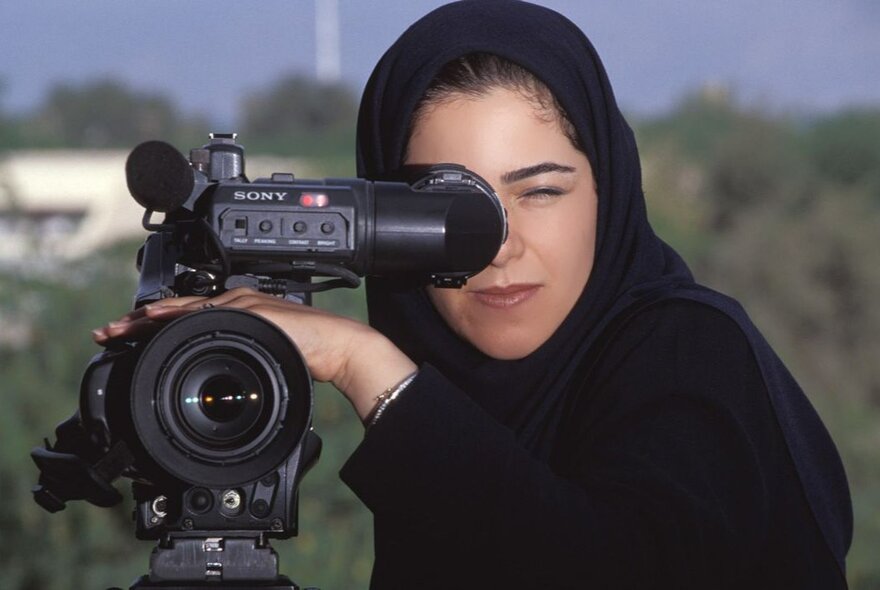 Iranian filmmaker Mozhgan Pazooki looking through a film camera lens, a landscape behind her.
