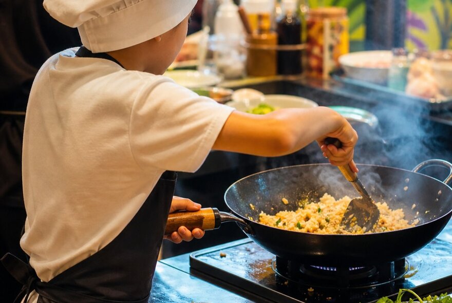 A child wearing a chef's hat and apron, stirring rice in a wok.