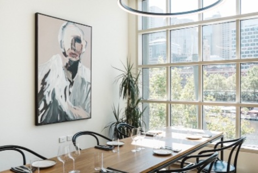 A sunlit private dining room in a wine bar, with large windows behind a timber table and chairs; pink-toned artwork on the wall.