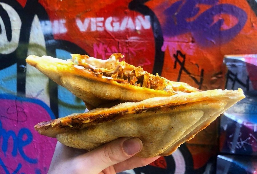 A hand holding a cheesy vegan jaffle in front of a colourful graffiti wall.