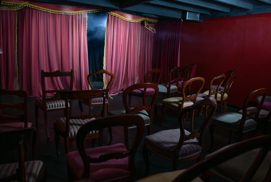 Small venue with curtained stage and vintage dining chairs.