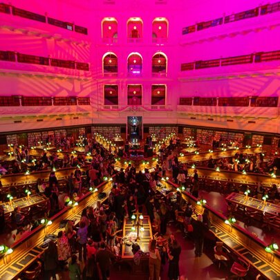 The Dome reading room of State Library Victoria showing the upper level balconies, and rows of desk fanning out from the centre, all bathed in a pink light.