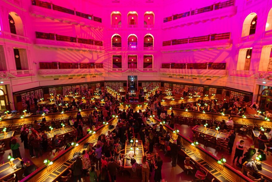 The Dome reading room of State Library Victoria showing the upper level balconies, and rows of desk fanning out from the centre, all bathed in a pink light.