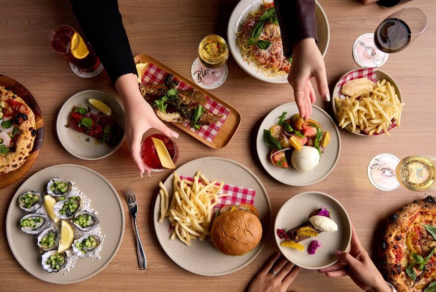 Looking down on a table set with many plates including burgers, fries and oysters, with hands reaching for dishes. 