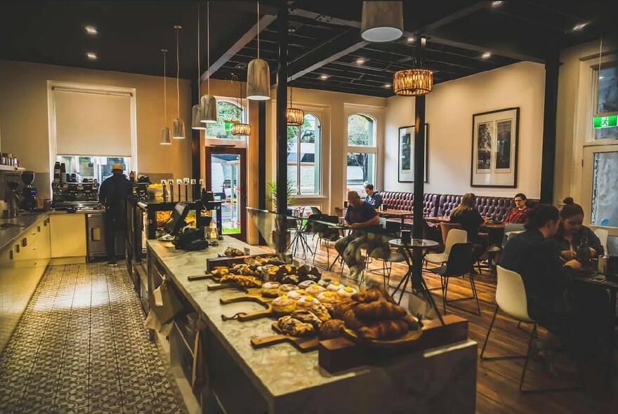 Cafe interior with banquettes, tables and chairs, and central counter with boards holding cakes and pastries.