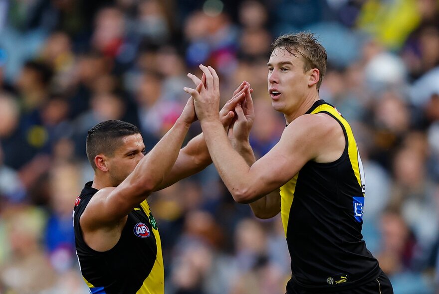 Two Richmond AFL players high-fiving and celebrating on the field, a blurred crowd in the stands behind them.