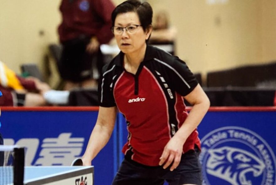 Table tennis coach Chau playing table tennis and concentrating with a paddle in her hand, standing behind the table.