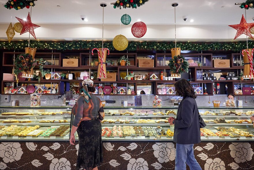 Two people browsing a cake counter with Christmas decorations hanging from the ceiling.