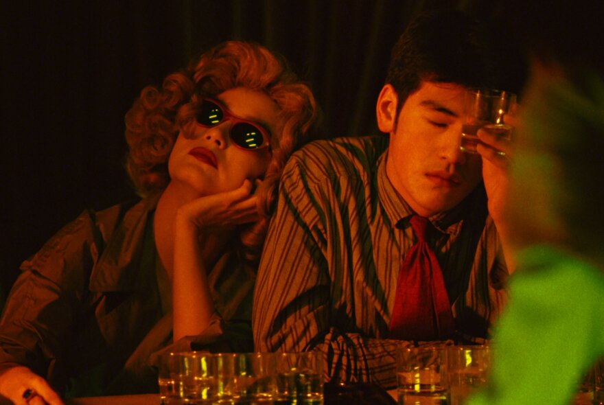 A still image from a film showing a darkened bar with two people drinking and looking exhausted, the woman wearing sunglasses and looking upwards. 