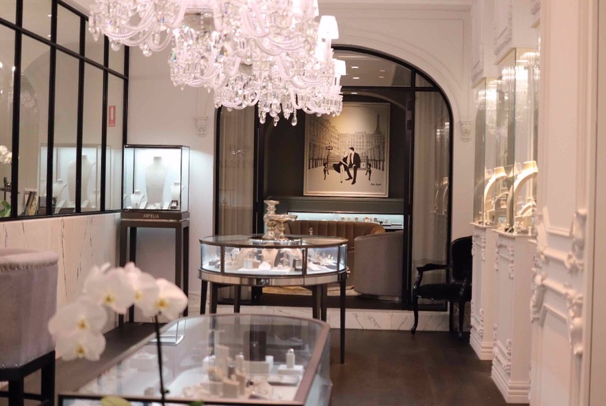 White-walled shop interior, with large painting at rear, display cases in middle, large chandelier from ceiling.