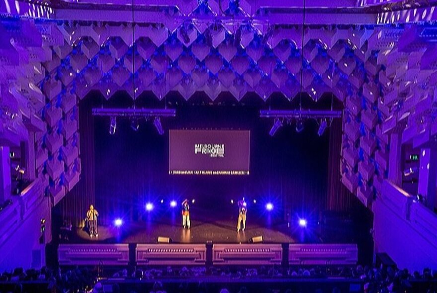 Three performers on stage at The Capitol Theatre, the art deco soundproofing lit up by purple lights.