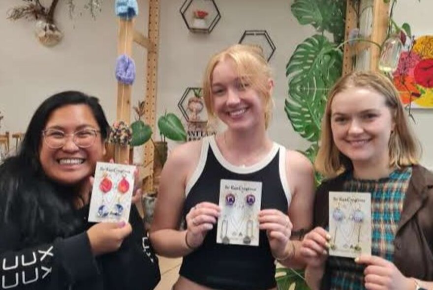 Three smiling workshop participants in a studio space, holding up jewellery they have made.