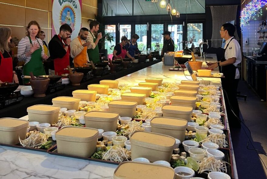 Long table inside a restaurant set up with cooking stations, ingredients and cooking utensils, with people on one side and a chef on the other side, ready for a Thai cooking workshop.