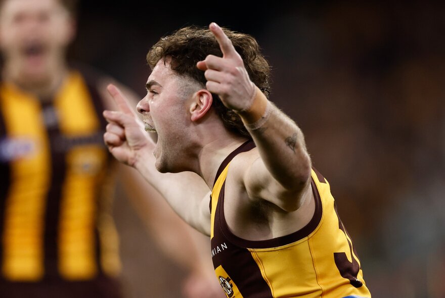 A Hawthorn player celebrates a goal with two fingers in the air, a blurred player in the background. 