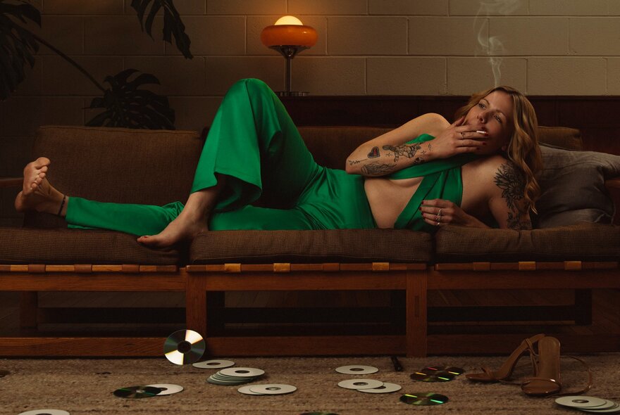 A woman in a green outfit lounging on a sofa, smoking a cigarette, with CDs strewn on the floor in front of her.