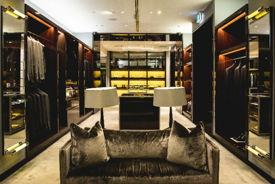 Mirrored walls on either side of a square room that includes a couch and racks of men's suits; interior of Harrolds department store.