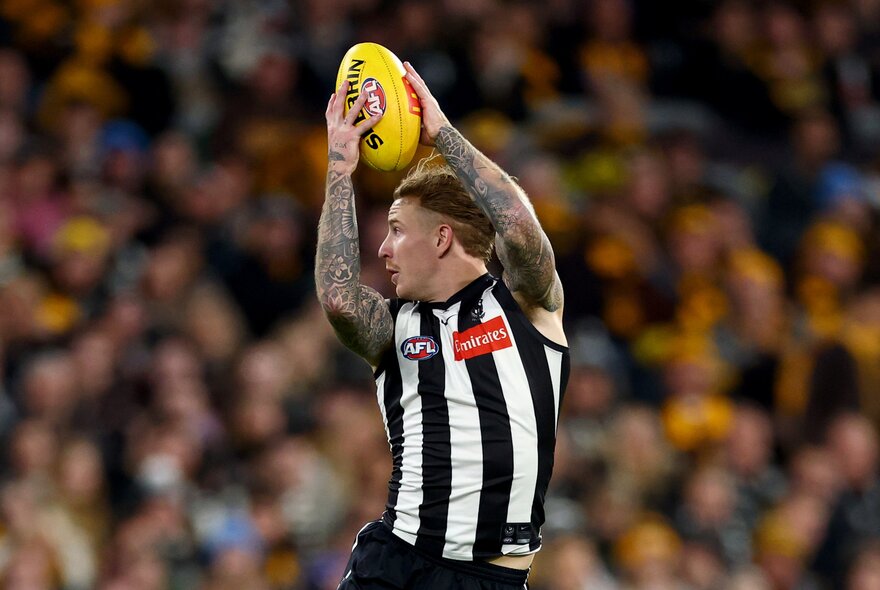 A Collingwood player marks a yellow football overhead during a match with a blurred crowd behind him. 