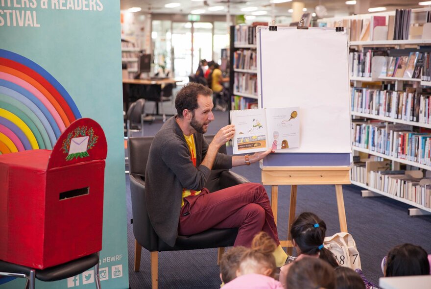 A bearded man seated in a chair holding a picture book in a library, children seated in front of him.