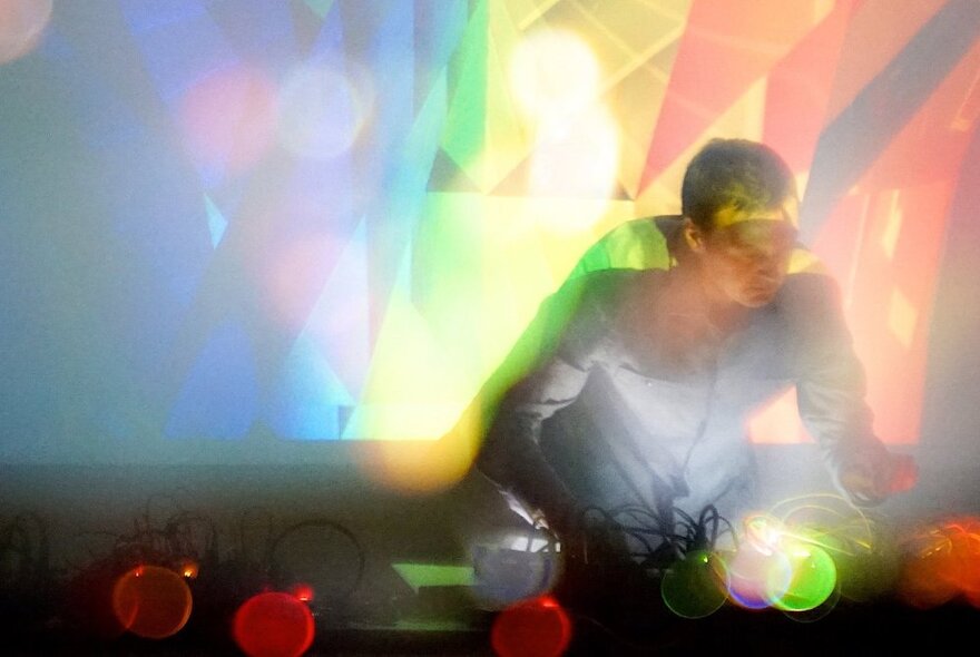 An out of focus person working at a mixing desk, with bright blurred yellow, green and orange lights surrounding them.