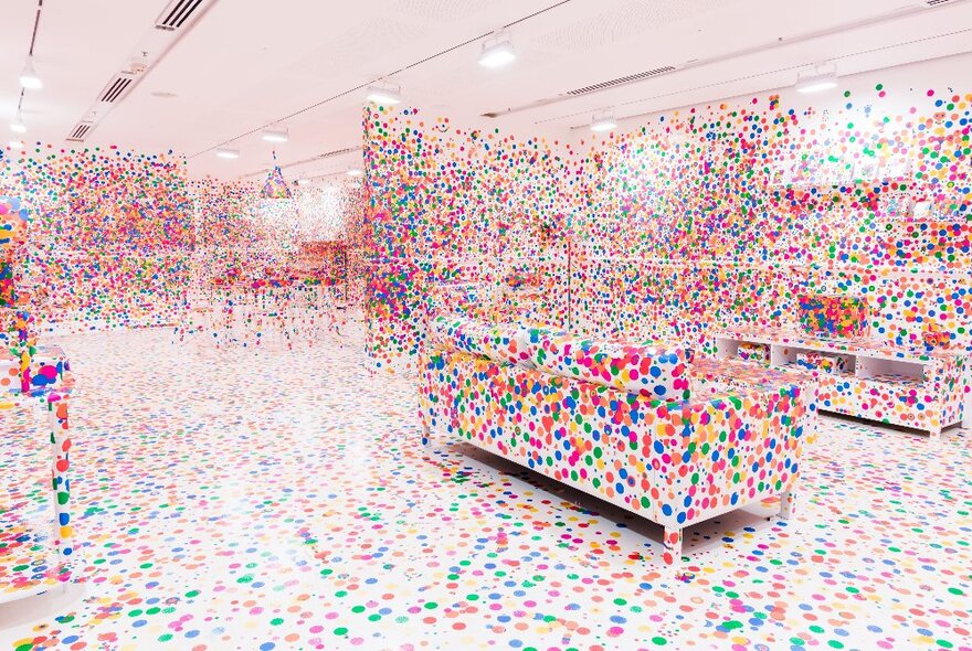 A white room with white floor, white walls and white furniture that is covered by brightly coloured stick-on dots.